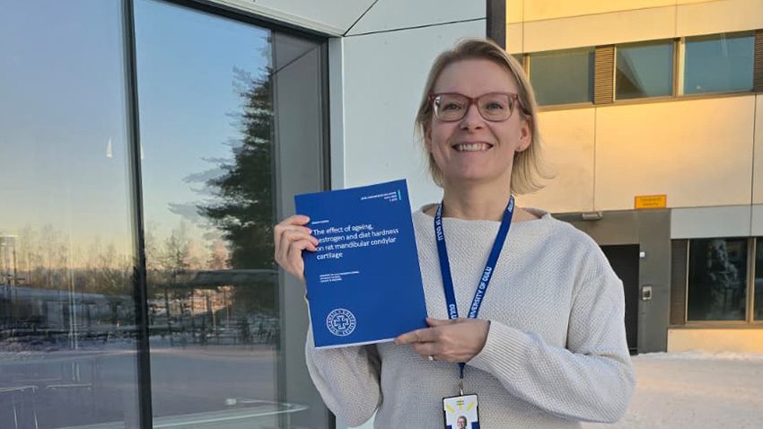 EHL Riikka Haurun väitös Väittelijän kädessä kirja, kuva pihalta rakennusten edestä.