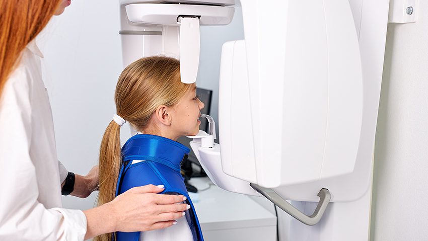 Säteilyfysiikka. Kuvassa lapssi kuvantamislaitteella ja hoitaja ohjaa häntä.